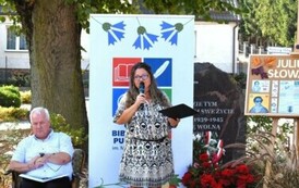 Przy rolce z logiem Biblioteki stoi Kierowniczka m&oacute;wiąc coś do mikrofonu. Z lewej strony siedzi Radny Gminy,a z prawej strony stoi tablica korkowa z informacjami o Juliuszu Słowackim.