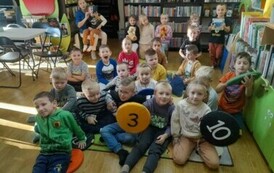 Dzieci siedzą na dywanie i słuchają bajki, kt&oacute;rą czyta im bibliotekarka. Dw&oacute;ch chłopc&oacute;w trzyma w rękach siedziska.