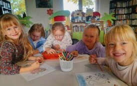 Przy białym stole siedzą uśmiechnięte dziewczynki i kolorują wiewi&oacute;rkę i jeżyka.