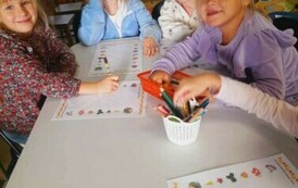 Przy białym stole siedzą dziewczynki i kredkami robią jesienne karty pracy.