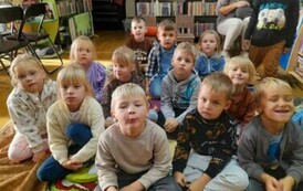 Dzieci siedzą na dywanie i słuchają bajki, kt&oacute;rą czyta im bibliotekarka.