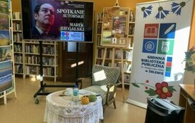 Po środku stoi okrągły stolik z fotelem. Po prawej stronie stoi rolap z logiem biblioteki, a po lewej stronie stoi duży ekran z wyświetlanym plakatem ze spotkania.