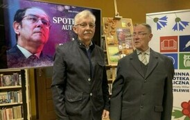 Pan Marek stoi z czytelnikiem. Za nimi stoi duży ekran z wyświetlonym plakatem ze spotkania. Z prawej strony stoi rolo z logiem biblioteki.