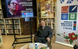 Autor wierszy siedzi przy stoliku na kt&oacute;rym jest kawa, ciasto i woda. Z prawej strony stoi rolo z logiem biblioteki,a z lewej strony stoi duży ekran z wyświetlonym plakatem o spotkaniu.