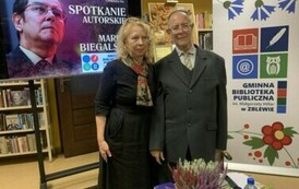 Autor wierszy stoi z czytelniczką przy stoliku. Po lewej stronie stoi duży kran z wyświetlonym plakatem o spotkaniu, a po prawej stronie stoi rolo z logiem biblioteki.