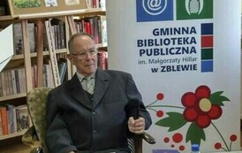 Autor wierszy siedzi przy stoliku na kt&oacute;rym jest kawa, ciasto i woda. Trzyma na kolanach ksera swoich wierszy,a w ręce trzyma mikrofon. Z prawej strony stoi rolo z logiem biblioteki.