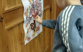 Na drzwiach jest przyklejony plakat z drzewem, przy nich stoi chłopiec i przykleja do drzewka listki.