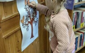 Na drzwiach jest przyklejony plakat z drzewem, przy nich stoi dziewczynka i przykleja do drzewka listki.