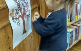 Na drzwiach jest przyklejony plakat z drzewem, przy nich stoi dziewczynka i przykleja do drzewka listki.