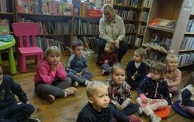 Dzieci siedzą na siedziskach w bibliotece wraz ze swoja Panią i słuchają czytanej bajki przez bibliotekarkę.