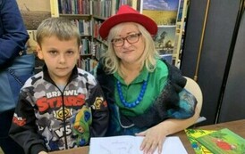 Chłopiec w jasnych włosach i w kolorowej bluzie stoi przy stoliku przy kt&oacute;rym Pani z teatrzyku podpisuje zakupione książki.