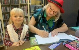 Dziewczynka w blond włosach w białej kamizelce stoi przy stoliu gdzie Pani z teatrzyku podpisuje dla niej książkę.