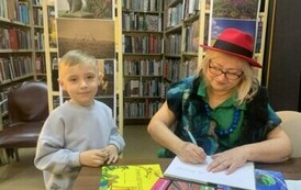 Chłopiec w blond włosach i w siwej bluzie stoi przy stoliku gdzie Pani z teatrzyku podpisuje dla niego książkę.