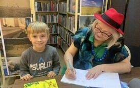 Chłopiec w blond włosach i w siwej bluzie stoi przy stoliku gdzie Pani z teatrzyku podpisuje dla niego książkę.