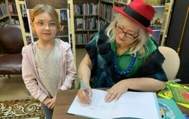 Dziewczynka w blond włosach w okularkach stoi przy stoliku gdzie Pani z teatrzyku podpisuje dla niej książkę.