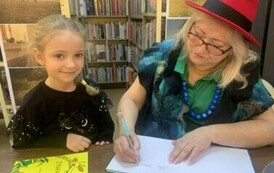 Dziewczynka w blond włosach i w czarnej bluzeczce stoi przy stoliku gdzie Pani z teatrzyku podpisuje dla niej książkę.