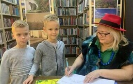 Dw&oacute;ch chłopc&oacute;w w blond włosach i w siwych bluzach stoją przy stoliku gdzie Pani z teatrzyku podpisuje dla nich książkę.