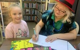 Dziewczynka w blond włosach i r&oacute;żowej bluzce stoi przy stoliu gdzie Pani z teatrzyku podpisuje dla niej książkę.