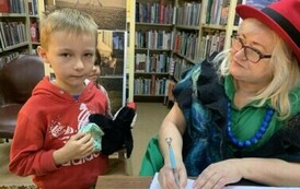 Chłopiec w jasnych włosach i w czerwonej bluzie z maskotką w ręku stoi przy stoliku gdzie Pani z teatrzyku podpisuje mu książkę.