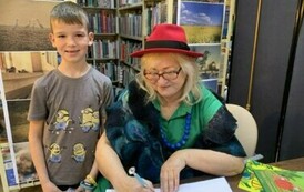 Chłopiec w jasnych włosach stoi przy stoliku gdzie siedzi Pani z teatrzyku i podpisuje mu książkę.