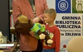 Pan w r&oacute;żowej marynarce i czerwonym kapeluszu trzyma leżącego misia pod brodą, a chłopiec stoi przy tym i się patrzy.