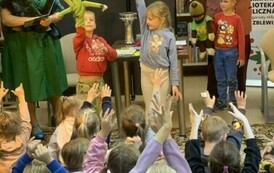 Dzieci siedzą na dywanie z podniesionymi rączkami, z przodu Pani w zielonej sukience coś czyta,a dwoje dzieci ma na ręce nałożone zielone duże łapki, a z prawej strony stoi Pan w czerwonym kapeluszu z podniesionymi rękoma,z chłopcem, kt&oacute;ry trzyma misia.