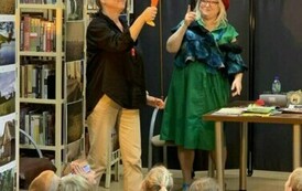 Dzieci siedzą na dywanie i patrzą na scenkę gdzie Pani w zielonej sukience pokazuje coś  palcem do g&oacute;ry,a Pani wychowaczyni trzyma u g&oacute;ry kukiełkę z dużym nosem.