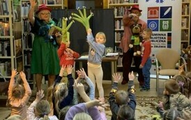 Dwoje dzieci stoi z przodu i mają ręce podniesione do g&oacute;ry i nałożone na jednej z nich zielone łapki, Pani z lewej strony coś czyta, a z prawej strony stoi chłopiec  przy Panu kt&oacute;ry trzyma misia.Dzieci z publicznośći mają ręce podniesione do g&oacute;ry.