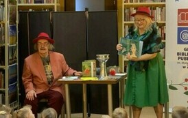 Dzieci siedzą i  oglądają scenkę gdzie przy stoliku siedzi Pan w czerwonym kapeluszu , obok stoi Pani w zielonej sukience i czerwonym kapeluszu pokazując otwartą książkę. Na stoliku stoją książki i puchar.