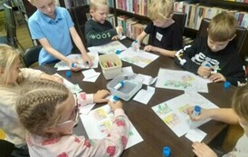 Dzieci siedzą przy stole i naklejają na białą kartkę części historyjki obrazkowej.