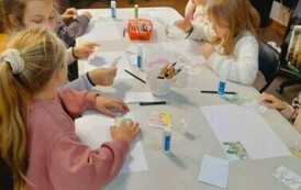 Dziewczynki przy białym stole przyklejają do białej kartki części układanki obrazkowej.