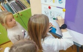 Dziewczynka w blond włosach przy tablicy  łączy lierki w wydrukowanych wagonach aby powstalo słowo "Przepraszam".Obok niej są inne dzieci.