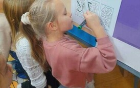 Dziewczynka w blond włosach w kitku  przy tablicy łączy literki w wydrukowanych wagonach aby powstało słowo "Dziękuję"