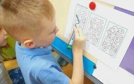 Chłopiec w blond włosach i niebieskiej koszulce przy tablicy łączy literki w wydrukowanych wagonach aby powstało słowo "proszę"