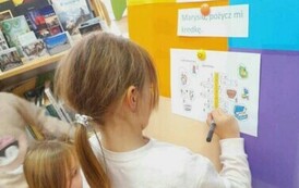 Dziewczynka w blond włosach w kitku wpisuje przy tablicy literki do krzyż&oacute;wki, obok niej widać inne dzieci. 