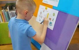 Chlopiec w blond włosach i niebieskiej koszulce stoi przy tablicy i uzupełnia literki w krzyż&oacute;wce.