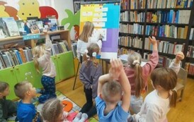 Dziewczynka w blond włosach stoi przy tablicy i uzupełnia literki w krzyż&oacute;wce,a za nią dzieci podnoszą ręce do g&oacute;ry zgłaszając chęć p&oacute;jścia do tablicy.