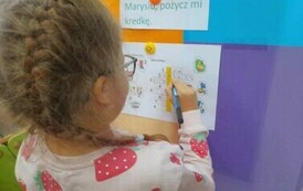 Dziewczynka w blond włosach w okularach przy tablicy wpisuje literki do krzyż&oacute;wki.