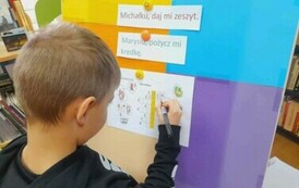 Chłopiec w blond włosach stoi przy tablicy i uzupełnia literkami krzyż&oacute;wkę.