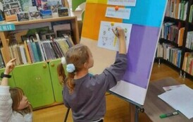 Dziewczynka klęczy przy tablicy i i wpisuje literki do krzyż&oacute;wki. Inna dziewczynka za nią podnosi rękę.