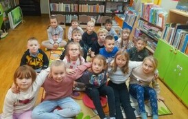Dzieci siedzą na dywanie i słuchają opowiadania, kt&oacute;re czyta bibliotekarka.