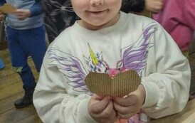 Dziewczynka w blond włosach pokazuje swoje serduszko wydarte ze starego papieru. W tle widac inne dzieci.