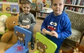 Dw&oacute;ch chłopc&oacute;w stoi przy dw&oacute;ch krzesełkach na kt&oacute;rych oparciach są naklejone głowy misi&oacute;w i przez otw&oacute;r w buzi misia dzieci ciągną nici kt&oacute;re leżą na krześle wizualizując jedzenie spagetti przez misia.