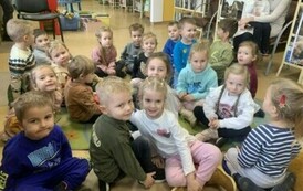 Dzieci siedzą na dywanie i słuchają bajki, kt&oacute;rą czyta im bibliotekarka.