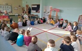 Dzieci siedzą w k&oacute;łku na sali i patrzą na bibliotekarkę, kt&oacute;ra pokazuje im plakat Mała Książka Wielki Człowiek. W sali obecni też są dwaj opiekunowie.