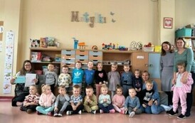 Dzieci siedzą w grupie na sali w szkole, a obok stoją dwie ich Panie i Pani bibliotekarka, kt&oacute;ra trzyma podziękowanie za zajęcia.