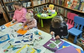 Dzieci stoją przy białym stole i wycinają z gazet misie i przyklejają je do kartki.