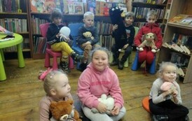 Dzieci siedzą na krzesełkach i siedziskach trzymając małe misie słuchają bajki.