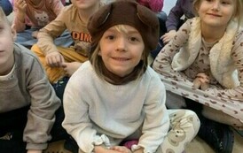 Dzieci siedzą na podłodze, jedno z dzieci z przodu ma ubrane misiową czapkę.