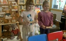 Dwie dziewczynki stoją z tyłu dw&oacute;ch krzeseł na kt&oacute;rych oparciach są naklejone głowy misi&oacute;w, a na siedziskach leżą talerzyki papierowe na kt&oacute;rych jest rozwinięta wł&oacute;czka. Dzieci ciągną wł&oacute;czkę, kt&oacute;ra przechodzi przez usta misia nawijając ją na rolkę wizual